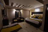 A cozy bedroom with modern decor features a large double bed adorned with yellow and black accented pillows and a throw. The room includes a wall-mounted television, a large mirror with lights on the sides, and a small seating area with two chairs and a table. There is also a small console table with a kettle and cups. The lighting is warm, enhancing the ambient feel of the room.