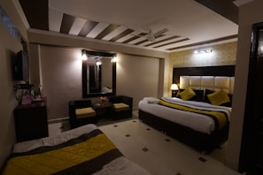 A cozy bedroom with modern decor features a large double bed adorned with yellow and black accented pillows and a throw. The room includes a wall-mounted television, a large mirror with lights on the sides, and a small seating area with two chairs and a table. There is also a small console table with a kettle and cups. The lighting is warm, enhancing the ambient feel of the room.