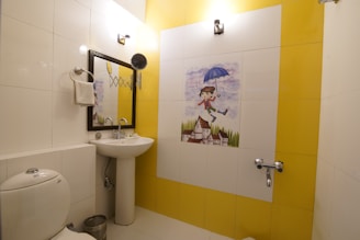 A bathroom with white and yellow tiled walls features a whimsical tile mural of a child holding an umbrella, floating above a village scene. There is a white ceramic toilet, a pedestal sink with a mirror above it, and a towel hanging on a ring. The room is well-lit with two wall-mounted light fixtures.