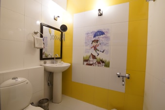 A bathroom with white and yellow tiled walls features a whimsical tile mural of a child holding an umbrella, floating above a village scene. There is a white ceramic toilet, a pedestal sink with a mirror above it, and a towel hanging on a ring. The room is well-lit with two wall-mounted light fixtures.