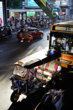 A busy city street scene features a colorful tuk-tuk and a public bus. The road is bustling with traffic, including motorbikes and cars. A large crowd of people is gathered on the sidewalk in front of shops and billboards. The architecture is modern, with a mix of commercial outdoor advertising and retail stores.