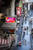 People walk along a bustling urban street next to a staircase leading to an elevated transport station. Brightly colored advertisements are visible on large screens and billboards. A person in uniform appears to be directing traffic, and various pedestrians pass by, some carrying bags. A taxi and motorcycles are present on the road to the left.