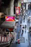 People walk along a bustling urban street next to a staircase leading to an elevated transport station. Brightly colored advertisements are visible on large screens and billboards. A person in uniform appears to be directing traffic, and various pedestrians pass by, some carrying bags. A taxi and motorcycles are present on the road to the left.
