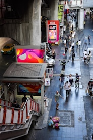 People walk along a bustling urban street next to a staircase leading to an elevated transport station. Brightly colored advertisements are visible on large screens and billboards. A person in uniform appears to be directing traffic, and various pedestrians pass by, some carrying bags. A taxi and motorcycles are present on the road to the left.