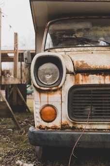The image depicts the front end of an old, rusted vehicle. The headlight is prominently visible, showing signs of age and corrosion. Rust stains streak down from the hood, with peeling paint and weathered metal. The setting appears to be outdoors, with grass and wooden structures partially visible in the background.