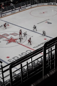 Ice hockey game in progress with players in red and white uniforms actively engaging on the rink. The ice is marked with lines and logos, and spectator seats are visible in the background.