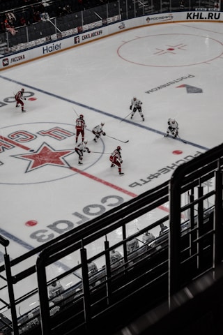Ice hockey game in progress with players in red and white uniforms actively engaging on the rink. The ice is marked with lines and logos, and spectator seats are visible in the background.