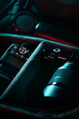 A camera bag containing various photography equipment, including a camera with a lens, a lens cap, and a flash. The items are organized in padded compartments with a soft interior lining. The lighting has a moody and professional tone, highlighting the equipment.