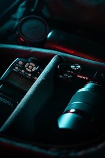 A camera bag containing various photography equipment, including a camera with a lens, a lens cap, and a flash. The items are organized in padded compartments with a soft interior lining. The lighting has a moody and professional tone, highlighting the equipment.