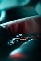A close-up view of a camera with the brand name Sony clearly visible. The image is stylized with a soft focus and lighting that emphasizes cool tones, creating an artistic effect. The focus is on the brand name and buttons of the camera, with a shallow depth of field.