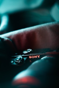 A close-up view of a camera with the brand name Sony clearly visible. The image is stylized with a soft focus and lighting that emphasizes cool tones, creating an artistic effect. The focus is on the brand name and buttons of the camera, with a shallow depth of field.