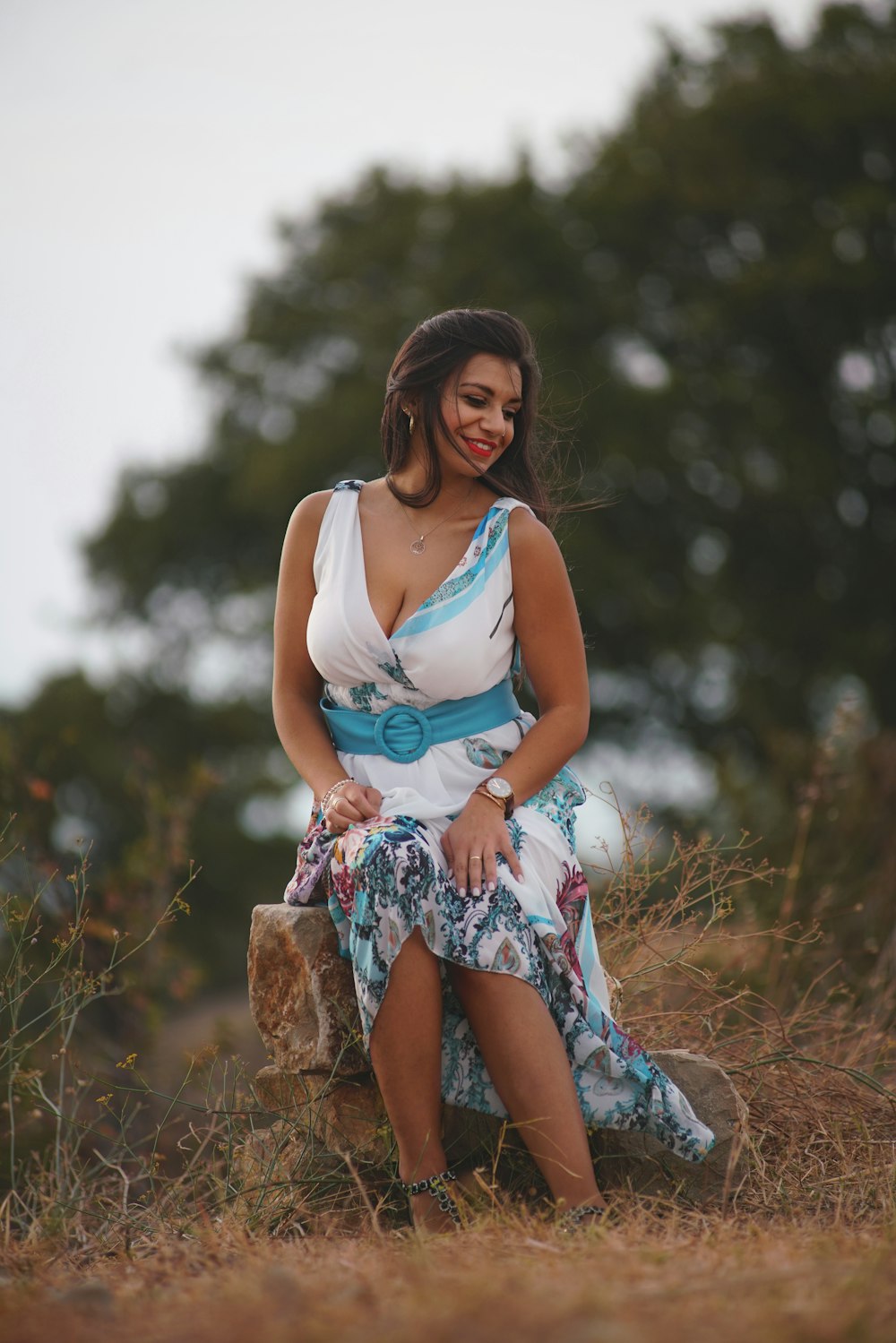 woman in white tank top and blue floral skirt sitting on brown rock during daytime