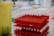 A stack of red racks filled with test tubes, each capped with a white lid, arranged in a clinical or laboratory setting. The background includes various laboratory equipment that is slightly out of focus.