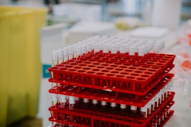 A stack of red racks filled with test tubes, each capped with a white lid, arranged in a clinical or laboratory setting. The background includes various laboratory equipment that is slightly out of focus.