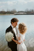 A couple stands close together by a serene lake, dressed in wedding attire. The groom, in a formal suit with a bowtie, holds a small bouquet of white flowers. The bride, in an elegant dress with lace sleeves, has curly hair adorned with a floral crown. The background features blurred trees and a cloudy sky.