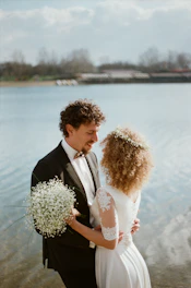 A couple stands close together by a serene lake, dressed in wedding attire. The groom, in a formal suit with a bowtie, holds a small bouquet of white flowers. The bride, in an elegant dress with lace sleeves, has curly hair adorned with a floral crown. The background features blurred trees and a cloudy sky.