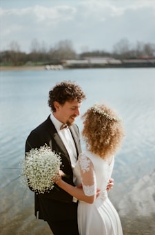 A couple stands close together by a serene lake, dressed in wedding attire. The groom, in a formal suit with a bowtie, holds a small bouquet of white flowers. The bride, in an elegant dress with lace sleeves, has curly hair adorned with a floral crown. The background features blurred trees and a cloudy sky.