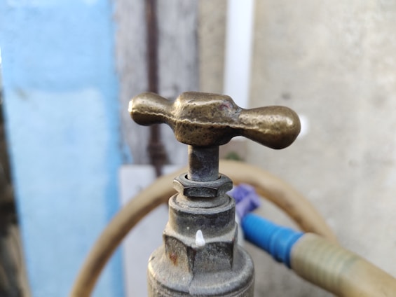 A close-up of a brass outdoor faucet with a patina, featuring a rounded handle and a background of a light blue wall and blurred hose.