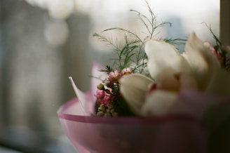 A delicate floral arrangement with pink and white flowers, possibly orchids and small pink blossoms, is wrapped in a light pink paper. Some green foliage is visible, providing a natural contrast to the pastel colors. The background is blurred, suggesting a window view that captures soft, diffused light.