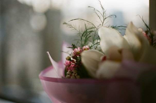 A delicate floral arrangement with pink and white flowers, possibly orchids and small pink blossoms, is wrapped in a light pink paper. Some green foliage is visible, providing a natural contrast to the pastel colors. The background is blurred, suggesting a window view that captures soft, diffused light.