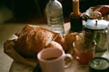 A table with various items including a loaf of bread, a smaller bread roll, a pink mug with a beverage, a glass of orange juice, a silver teapot, a tall water bottle, a yellow lit candle in a planter-like holder, and a bowl with tomatoes and flowers.