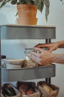 A person is organizing a shelf with glass jars filled with grains and oats. There's a pot with a green plant on the top shelf. Below, paper bags contain vegetables.