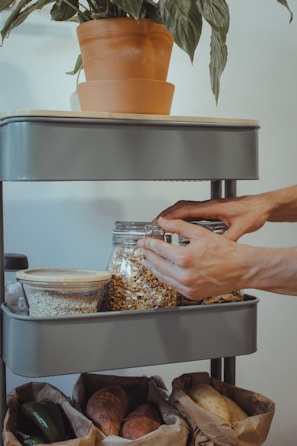 A person is organizing a shelf with glass jars filled with grains and oats. There's a pot with a green plant on the top shelf. Below, paper bags contain vegetables.