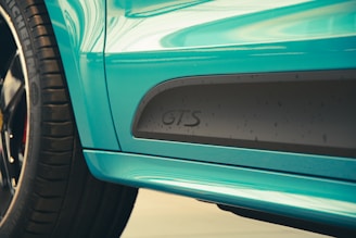 Close-up view of a turquoise-colored car with the letters 'GTS' displayed on the side near the wheel. The car features a glossy finish, and the tire with distinct tread patterns is visible on the left. Lighting emphasizes the reflective surface and the contours of the vehicle's body.