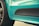 Close-up view of a turquoise-colored car with the letters 'GTS' displayed on the side near the wheel. The car features a glossy finish, and the tire with distinct tread patterns is visible on the left. Lighting emphasizes the reflective surface and the contours of the vehicle's body.