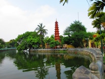 A serene landscape featuring a picturesque lake surrounded by lush green trees. A prominent red pagoda stands in the background, adding a touch of cultural architecture. The reflective water captures the surrounding scenery, creating a tranquil ambiance.