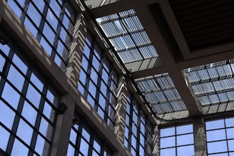 Sunlight streams through a series of large, grid-patterned glass windows, casting intricate shadow patterns on the interior walls. The design gives a modern and geometric aesthetic, with a mix of light and shadow creating a dynamic visual effect.
