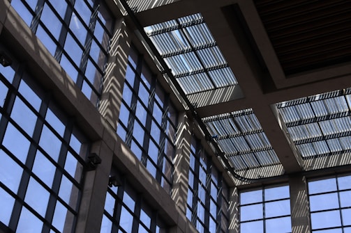 Sunlight streams through a series of large, grid-patterned glass windows, casting intricate shadow patterns on the interior walls. The design gives a modern and geometric aesthetic, with a mix of light and shadow creating a dynamic visual effect.