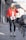 A fashionable person walks down an urban street. They are wearing a vibrant red jacket, black leather-like leggings, white boots with black laces, and a pair of earmuffs. They also carry a stylish white shoulder bag with a heart design. The background features parked cars and urban architecture.