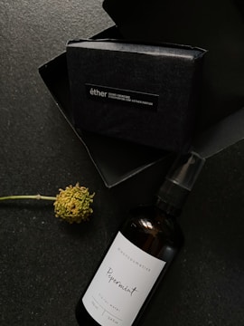 A dark packaging box labeled 'éther' is accompanied by a dark brown spray bottle labeled 'dixoncosmetics Peppermint' next to a single flower with a green stem.