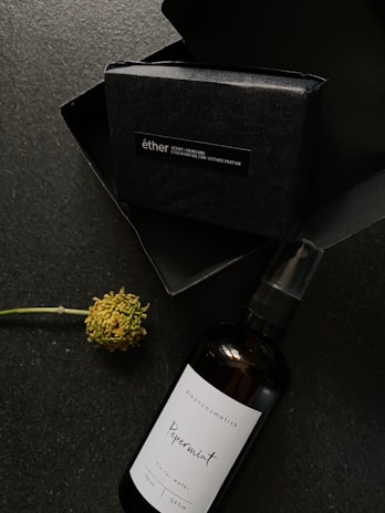 A dark packaging box labeled 'éther' is accompanied by a dark brown spray bottle labeled 'dixoncosmetics Peppermint' next to a single flower with a green stem.