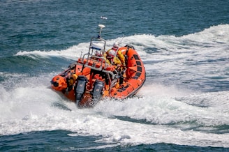 An orange inflatable rescue boat speeds through choppy waves, leaving behind a trail of foamy water. Several people wearing yellow life jackets and helmets are on board, indicating a focus on safety. The boat is equipped with twin outboard motors and has various equipment mounted on it.
