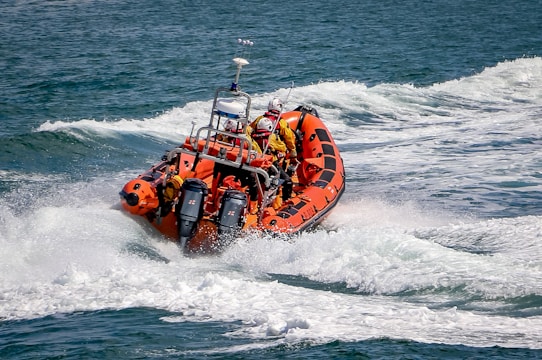 An orange inflatable rescue boat speeds through choppy waves, leaving behind a trail of foamy water. Several people wearing yellow life jackets and helmets are on board, indicating a focus on safety. The boat is equipped with twin outboard motors and has various equipment mounted on it.