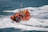 An orange inflatable rescue boat speeds through choppy waves, leaving behind a trail of foamy water. Several people wearing yellow life jackets and helmets are on board, indicating a focus on safety. The boat is equipped with twin outboard motors and has various equipment mounted on it.