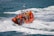 An orange inflatable rescue boat speeds through choppy waves, leaving behind a trail of foamy water. Several people wearing yellow life jackets and helmets are on board, indicating a focus on safety. The boat is equipped with twin outboard motors and has various equipment mounted on it.
