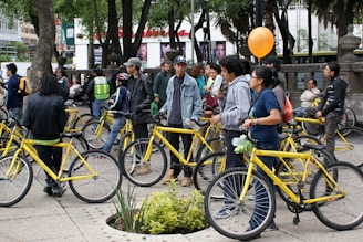 A group of people gather outdoors with yellow bicycles, standing and chatting in a tree-filled urban area. Some individuals are holding or wearing helmets, and one holds an orange balloon. The environment suggests a casual, social gathering or community event involving bicycles.