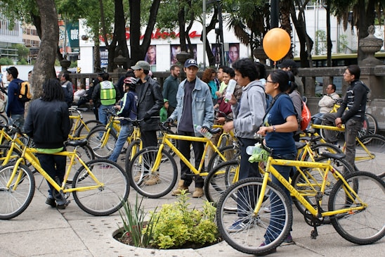 A group of people gather outdoors with yellow bicycles, standing and chatting in a tree-filled urban area. Some individuals are holding or wearing helmets, and one holds an orange balloon. The environment suggests a casual, social gathering or community event involving bicycles.