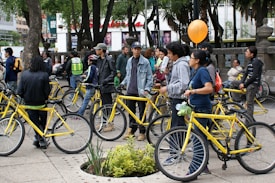A group of people gather outdoors with yellow bicycles, standing and chatting in a tree-filled urban area. Some individuals are holding or wearing helmets, and one holds an orange balloon. The environment suggests a casual, social gathering or community event involving bicycles.