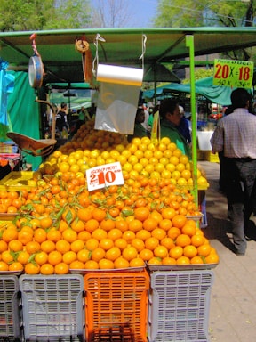 A vibrant outdoor market stall displaying an abundance of oranges stacked in crates. The sign with pricing information is prominent among the fruit. There is a canopy providing shade, and a few people are visible in the background.