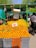 A vibrant outdoor market stall displaying an abundance of oranges stacked in crates. The sign with pricing information is prominent among the fruit. There is a canopy providing shade, and a few people are visible in the background.