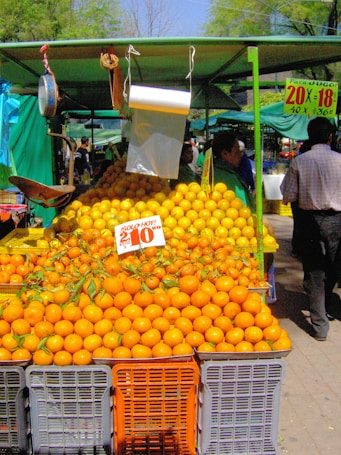 A vibrant outdoor market stall displaying an abundance of oranges stacked in crates. The sign with pricing information is prominent among the fruit. There is a canopy providing shade, and a few people are visible in the background.