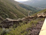 Wooden boardwalk winding through a lush, green valley, surrounded by dense vegetation and rugged hills. The path offers an elevated view of the natural landscape, providing a scenic route for walking.