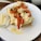 A white plate features a piece of toasted bread topped with a creamy spread, roasted red peppers, and almond slices. A small glass bowl containing cheese sticks is placed on the side. Drops of olive oil and herbs are also visible on the plate.