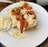 A white plate features a piece of toasted bread topped with a creamy spread, roasted red peppers, and almond slices. A small glass bowl containing cheese sticks is placed on the side. Drops of olive oil and herbs are also visible on the plate.
