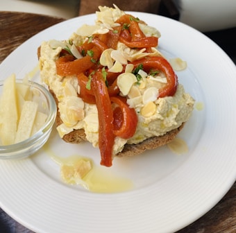 A white plate features a piece of toasted bread topped with a creamy spread, roasted red peppers, and almond slices. A small glass bowl containing cheese sticks is placed on the side. Drops of olive oil and herbs are also visible on the plate.