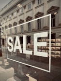 A storefront window displays the word 'SALE' in large white letters. The reflection on the glass shows a street scene with an old, classical-style building, complete with ornate windows. Inside the window, a variety of shoes are organized in a neat display.